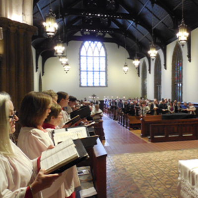 A church choir in white robes sings from hymnals at the front of a large, traditional church, while the congregation sits in pews facing a stained-glass window.