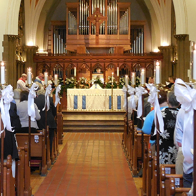A church interior with people seated in pews facing an altar decorated with flowers and white bows on the aisle. An organ and arched ceiling are visible in the background.
