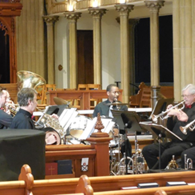 A brass quintet performs inside a church, surrounded by wooden pews and tall columns. Five musicians play brass instruments, including trumpet, trombone, and tuba, with sheet music on stands in front of them.