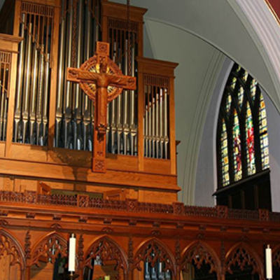A large wooden pipe organ and a crucifix inside a church, with arched ceilings and a stained glass window visible to the right.
