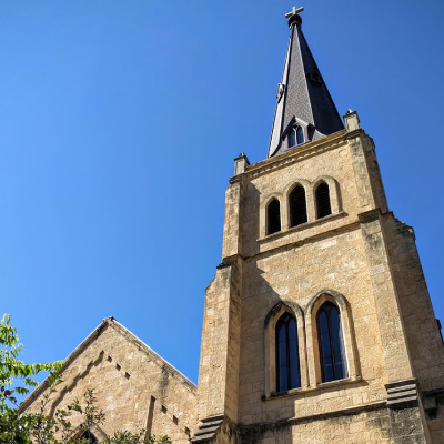 A tall stone church with arched windows and a pointed steeple topped by a cross rises against a clear blue sky, with some green tree branches visible in the lower left corner.