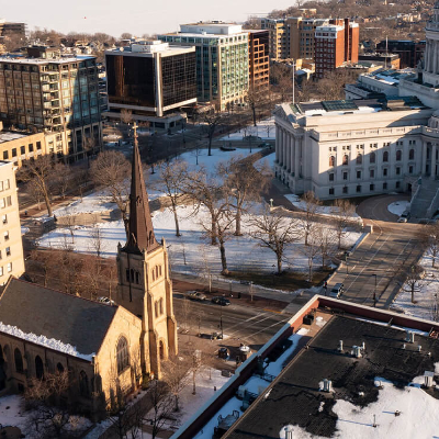 Aerial view of a cityscape with a stone church featuring a tall steeple, surrounded by leafless trees and snow-dusted ground, modern office buildings, and a large white government building in the background.